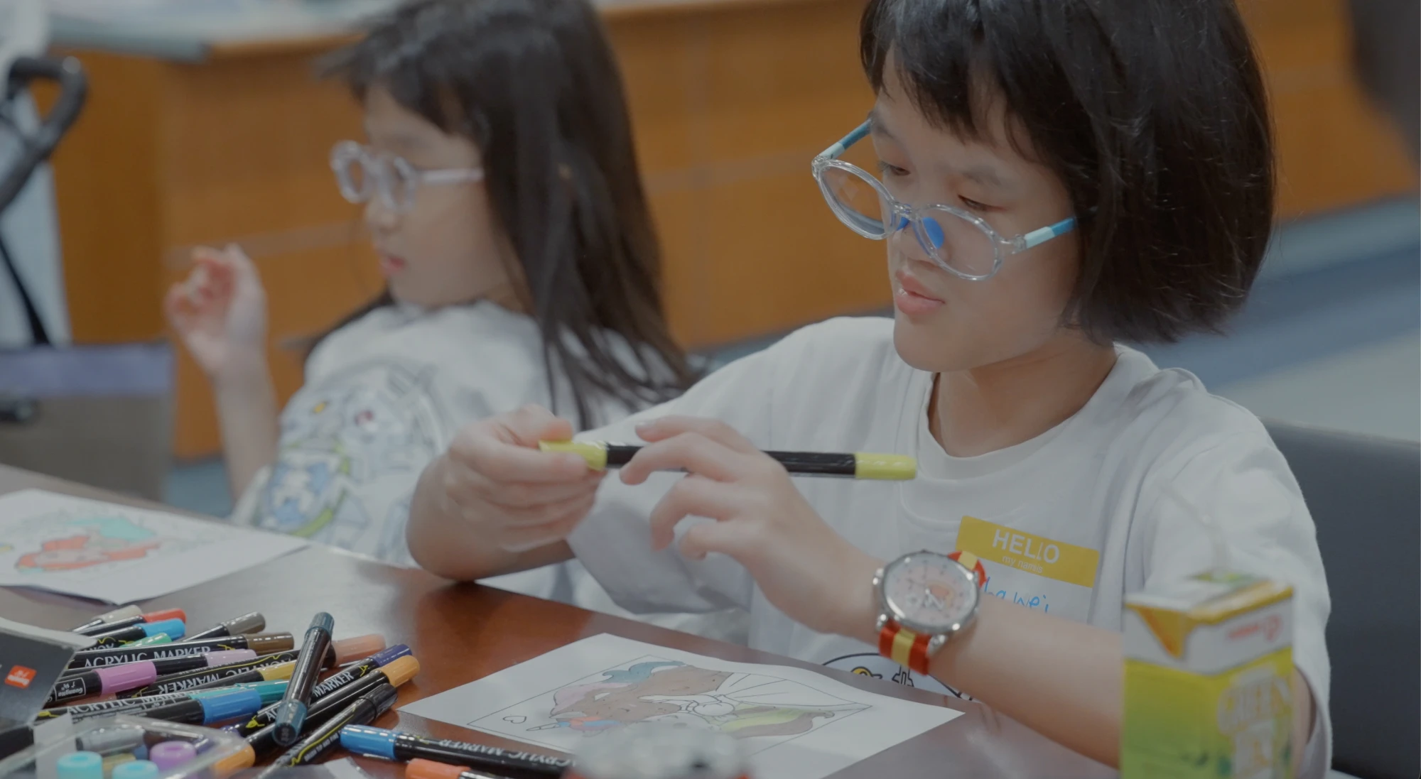 Two children in white Undiagnosed Hackathon T-shirts sit at a table colouring pictures with acrylic markers during the Undiagnosed Hackathon